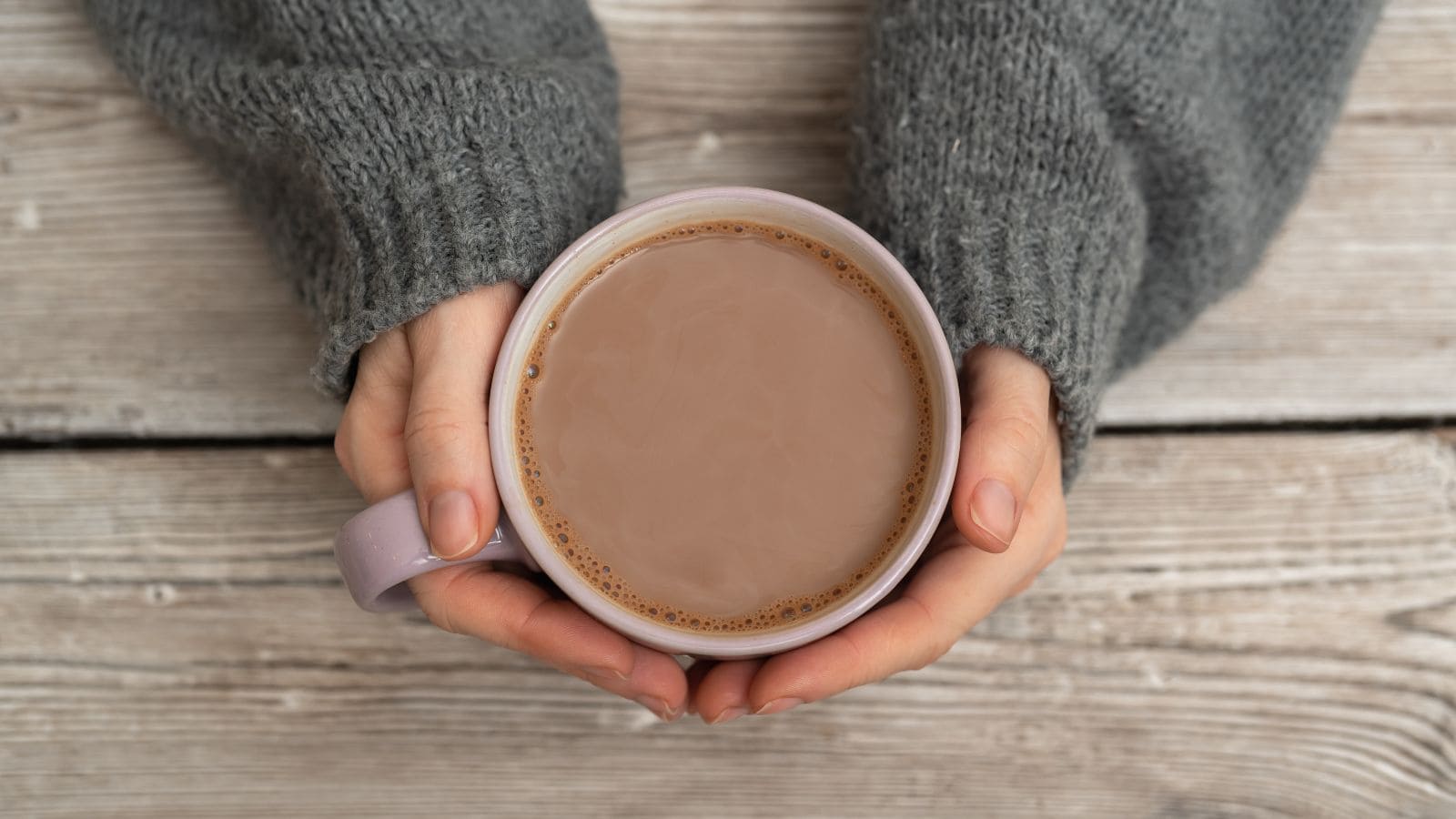 A person wearing a gray sweater holds a mug filled with light brown coffee or tea, viewed from above, on a wooden surface.