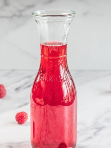 A clear glass carafe filled with red liquid is placed on a white marble surface. Four raspberries are scattered around the base of the carafe. The background is a white marble wall.