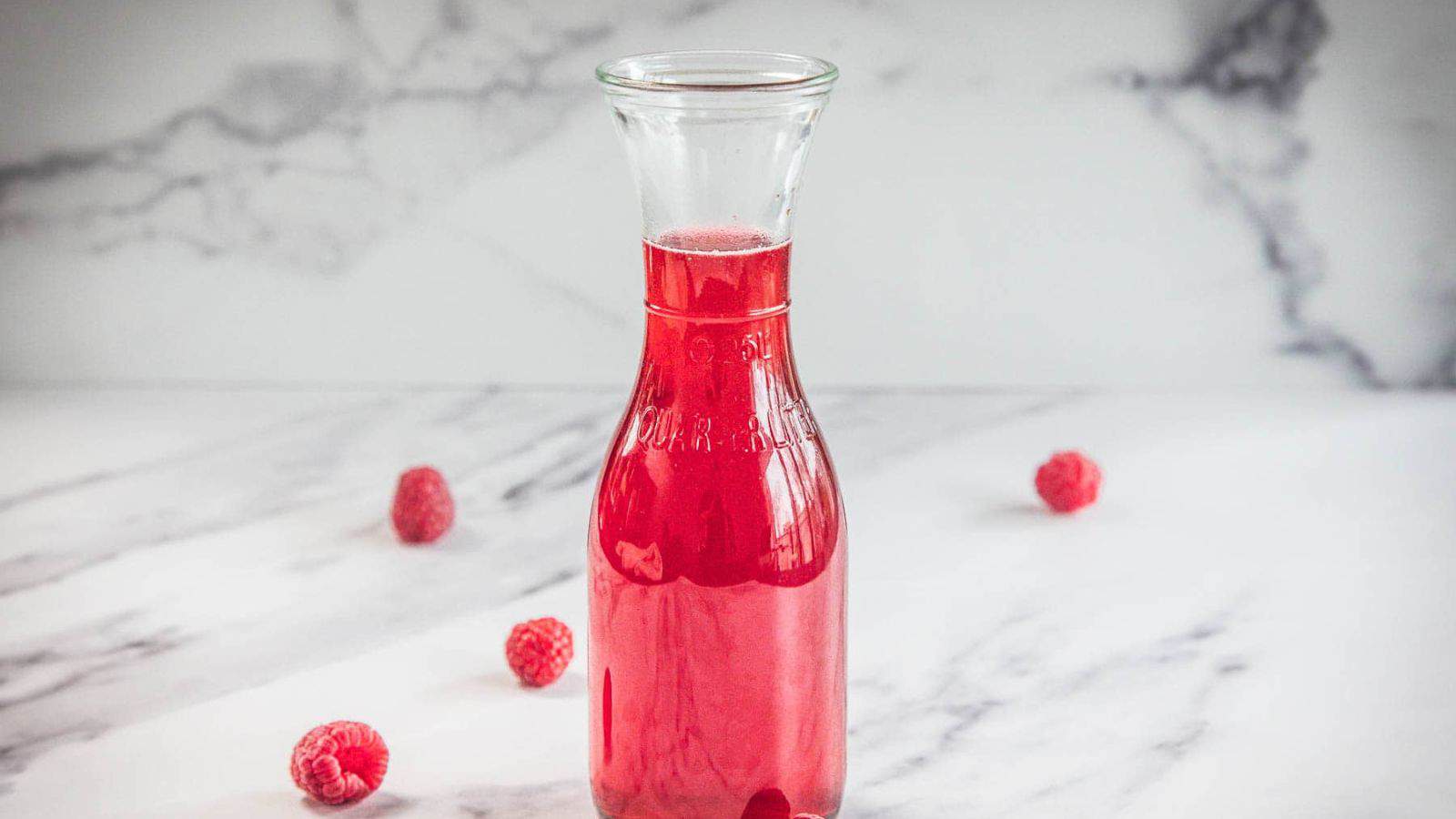 A clear glass carafe filled with red liquid is placed on a white marble surface. Four raspberries are scattered around the base of the carafe. The background is a white marble wall.