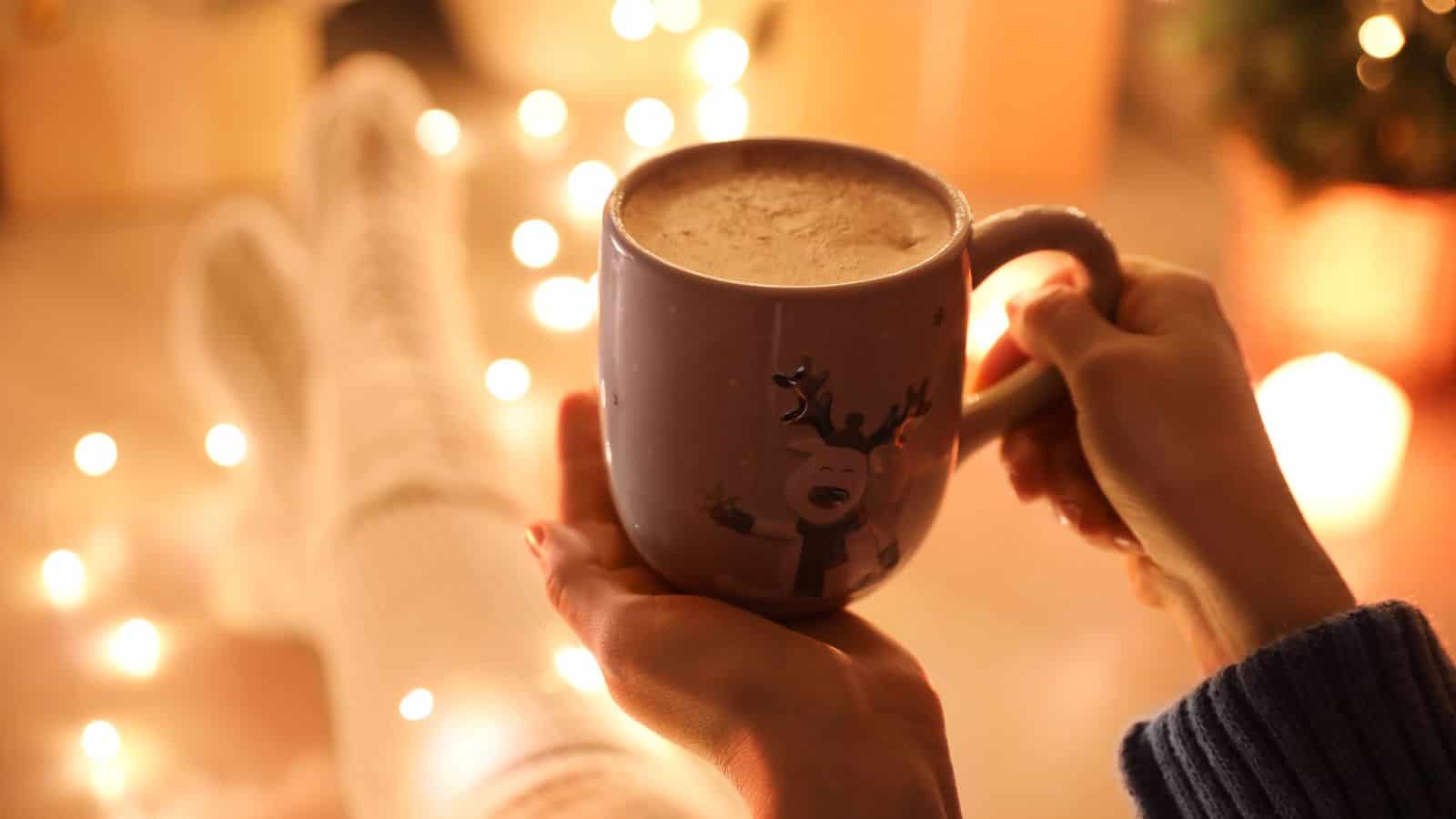 A person wearing cozy white socks holds a mug with a reindeer design, surrounded by warm string lights and a softly lit background.