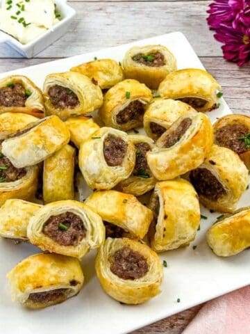 A square white plate holds several golden-brown sausage rolls, garnished with chopped herbs. A square dish of white dipping sauce and a bouquet of purple flowers are in the background on a wooden table.