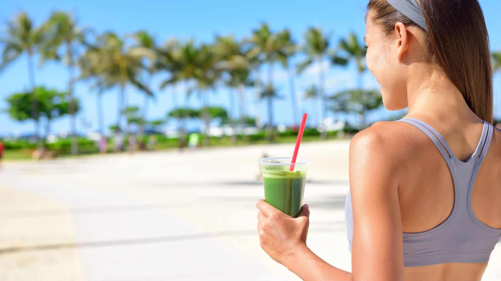 A woman in athletic wear stands outdoors holding a green smoothie with a red straw. Palm trees and a clear blue sky are visible in the background.