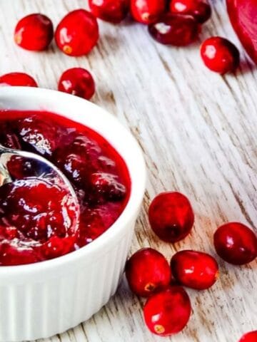 A white ramekin filled with cranberry sauce and a spoon, surrounded by whole cranberries and an orange, sits on a light wooden surface. A glass jar of cranberry sauce is visible in the background.