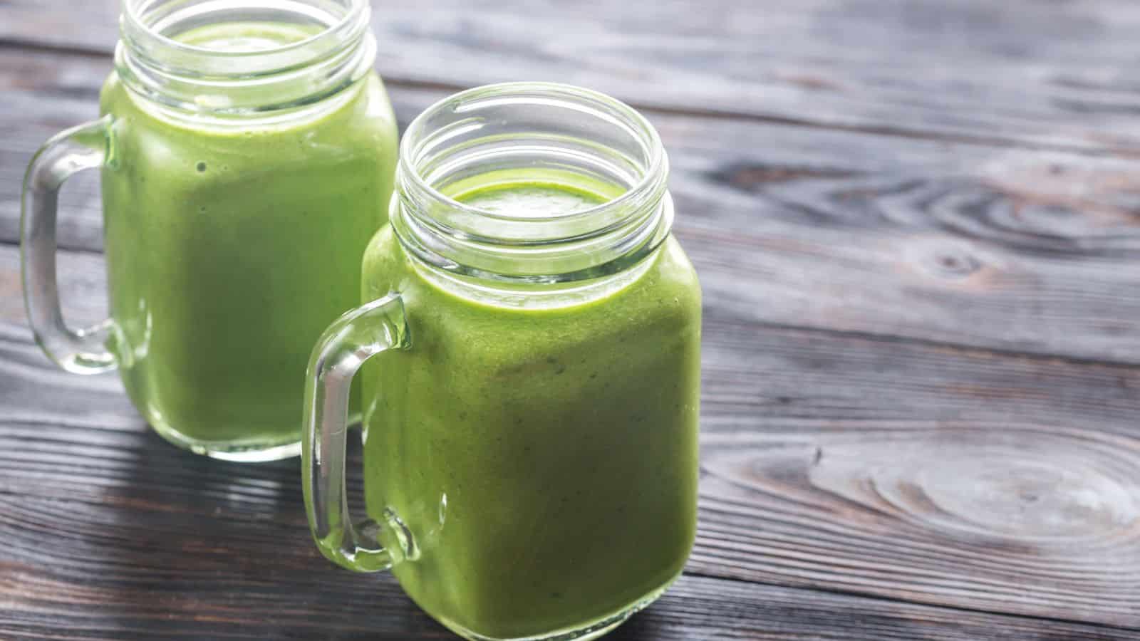 Two glass mason jar mugs with handles, each filled with a thick green smoothie, are placed on a wooden surface. The smoothie appears creamy and the jars are positioned close together.