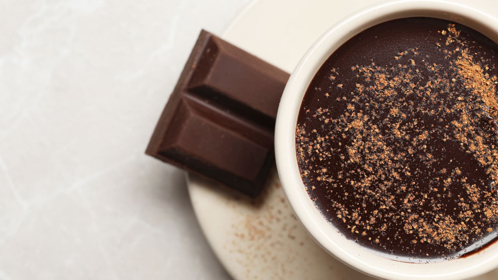 A cup of thick hot chocolate sprinkled with cocoa powder sits on a white plate next to two pieces of dark chocolate on a light-colored surface.