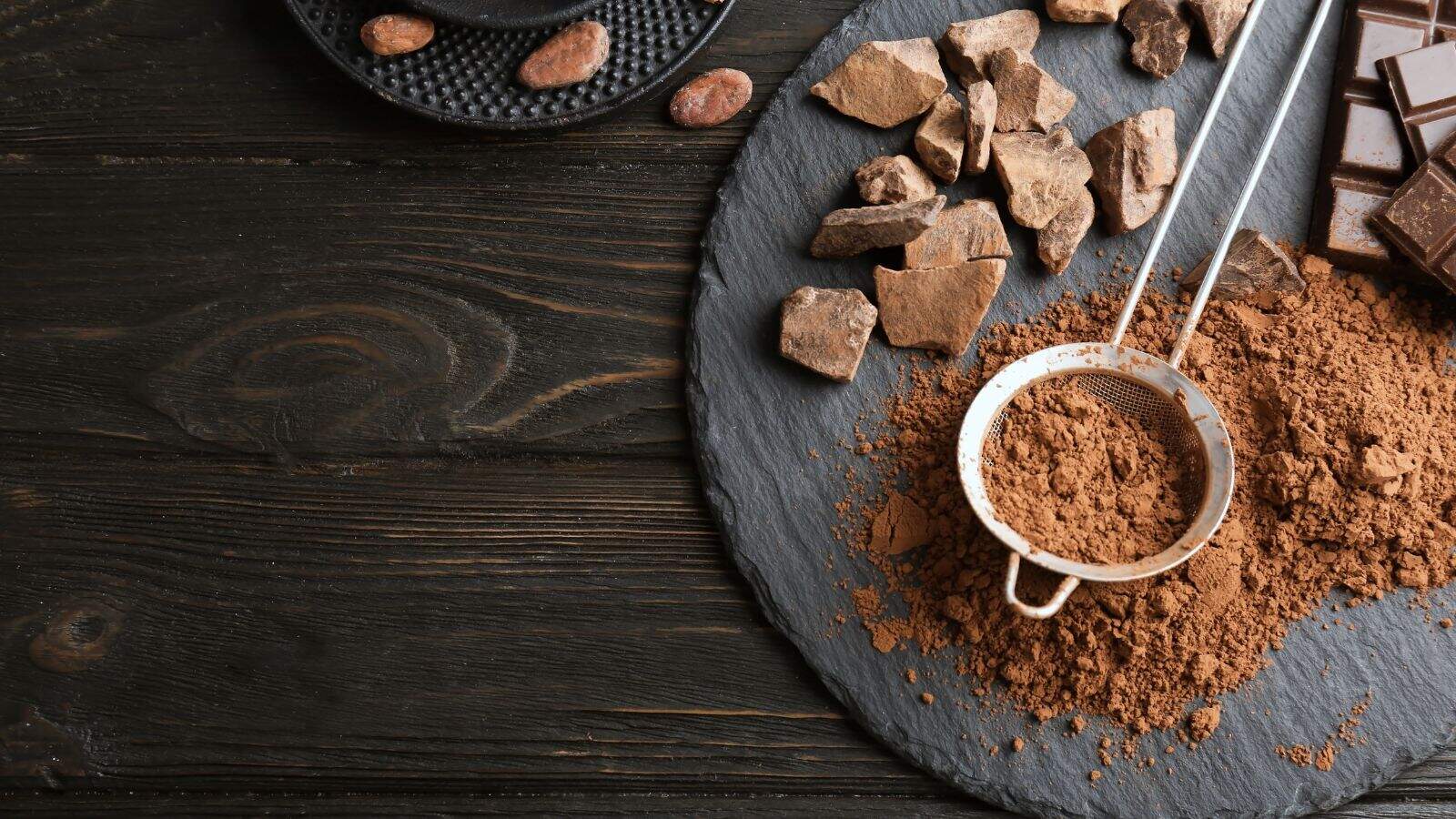 A metal sifter filled with cocoa powder rests on a slate board with cocoa powder, chocolate chunks, and cocoa beans on a dark wooden table.