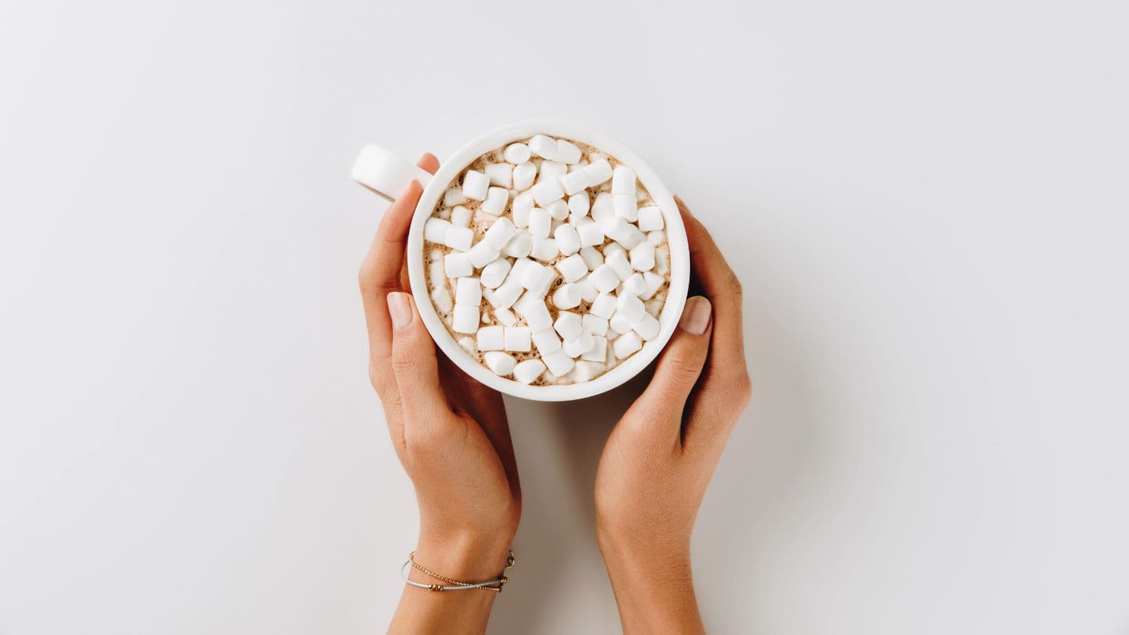 A pair of hands holds a white mug filled with hot chocolate topped with mini marshmallows, photographed from above against a white background.