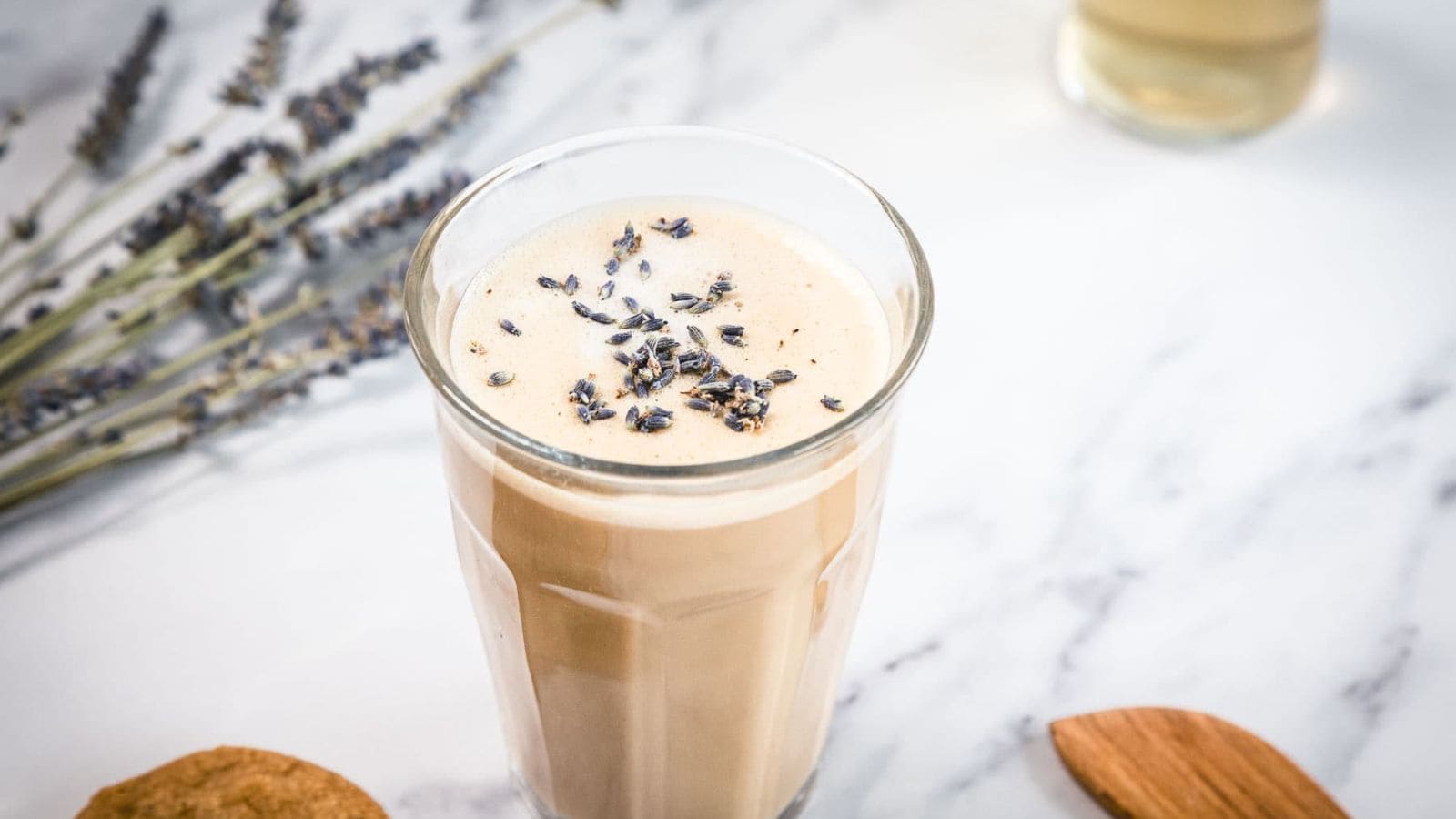 A glass of frothy iced coffee topped with dried lavender buds sits on a white marble surface. Lavender sprigs, a wooden spoon, and a cookie are nearby.