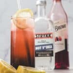 A tall glass of red 4th of July lemonade cocktail with lemon slices, next to bottles of non-alcoholic vodka and grenadine.
