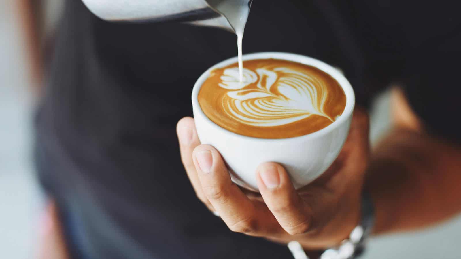 A person pours steamed milk to create latte art on a cup of coffee held in their hand, showcasing a moment from a guide to different types of coffee.