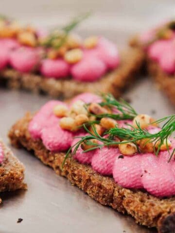 Four rectangular pieces of brown bread topped with pink spread, toasted pine nuts, and fresh dill are arranged on a metal tray.