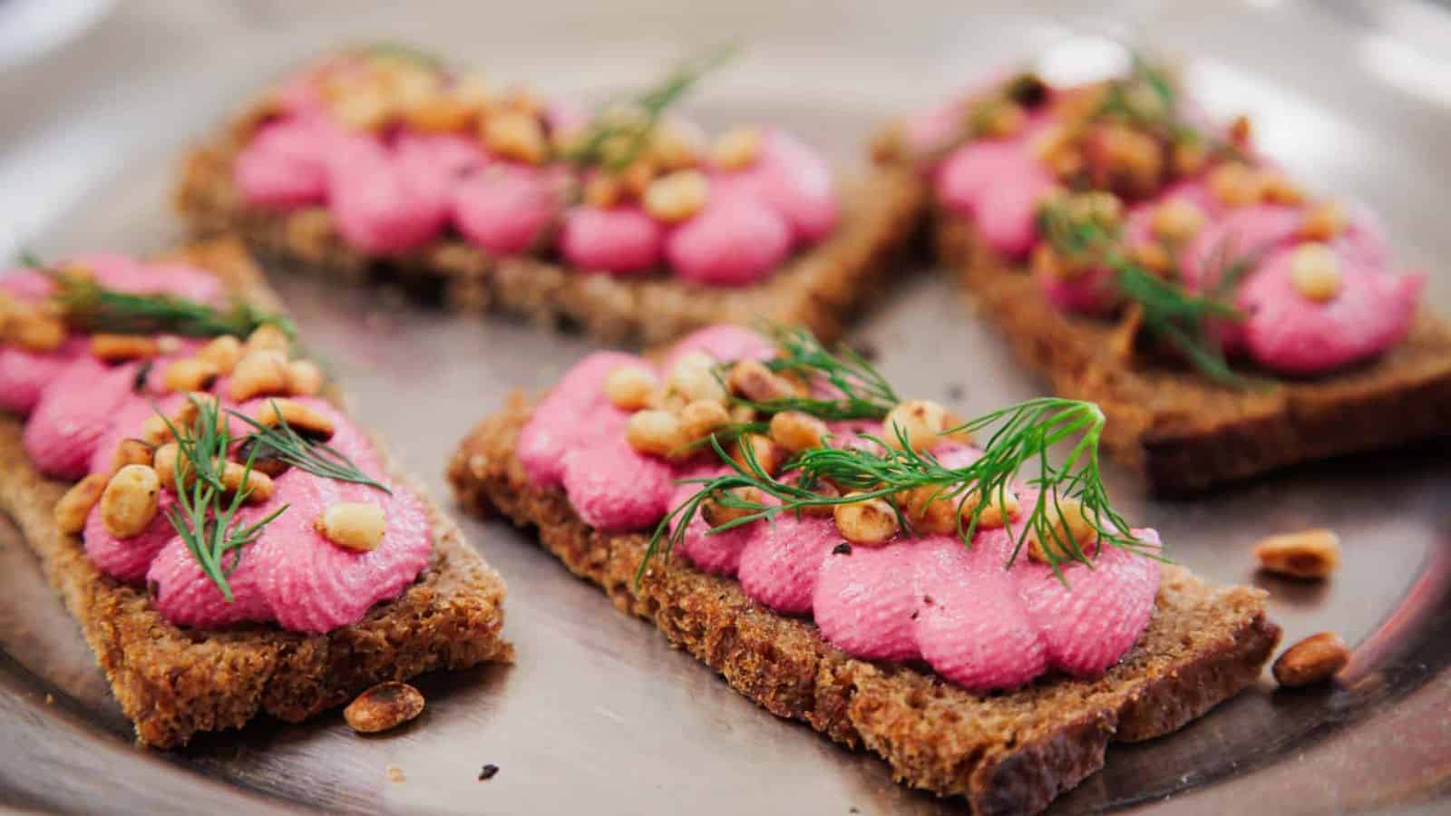 Four rectangular pieces of brown bread topped with pink spread, toasted pine nuts, and fresh dill are arranged on a metal tray.