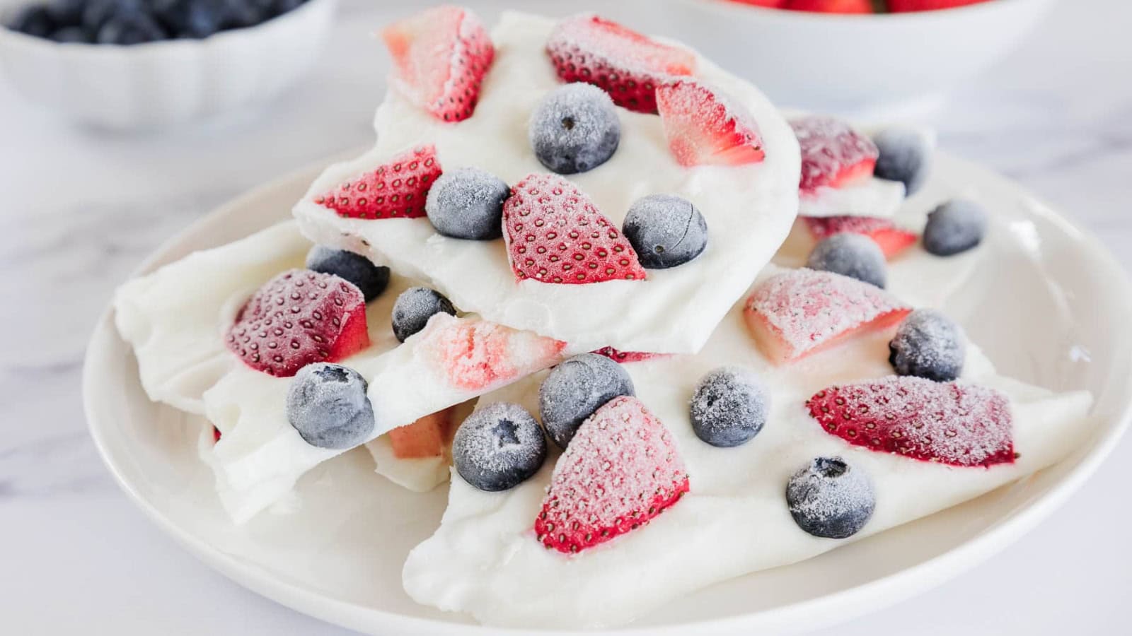 A plate of frozen yogurt bark topped with sliced strawberries and whole blueberries. The bark pieces are white with visible frost, and bowls of additional berries are blurred in the background.