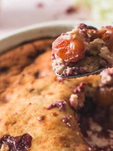 A baked fruit cobbler in a round white dish with a portion already served. A spoon holds up a serving, showing fruit filling and golden-brown crust. Cherries and crumbs are visible on the table in the background.