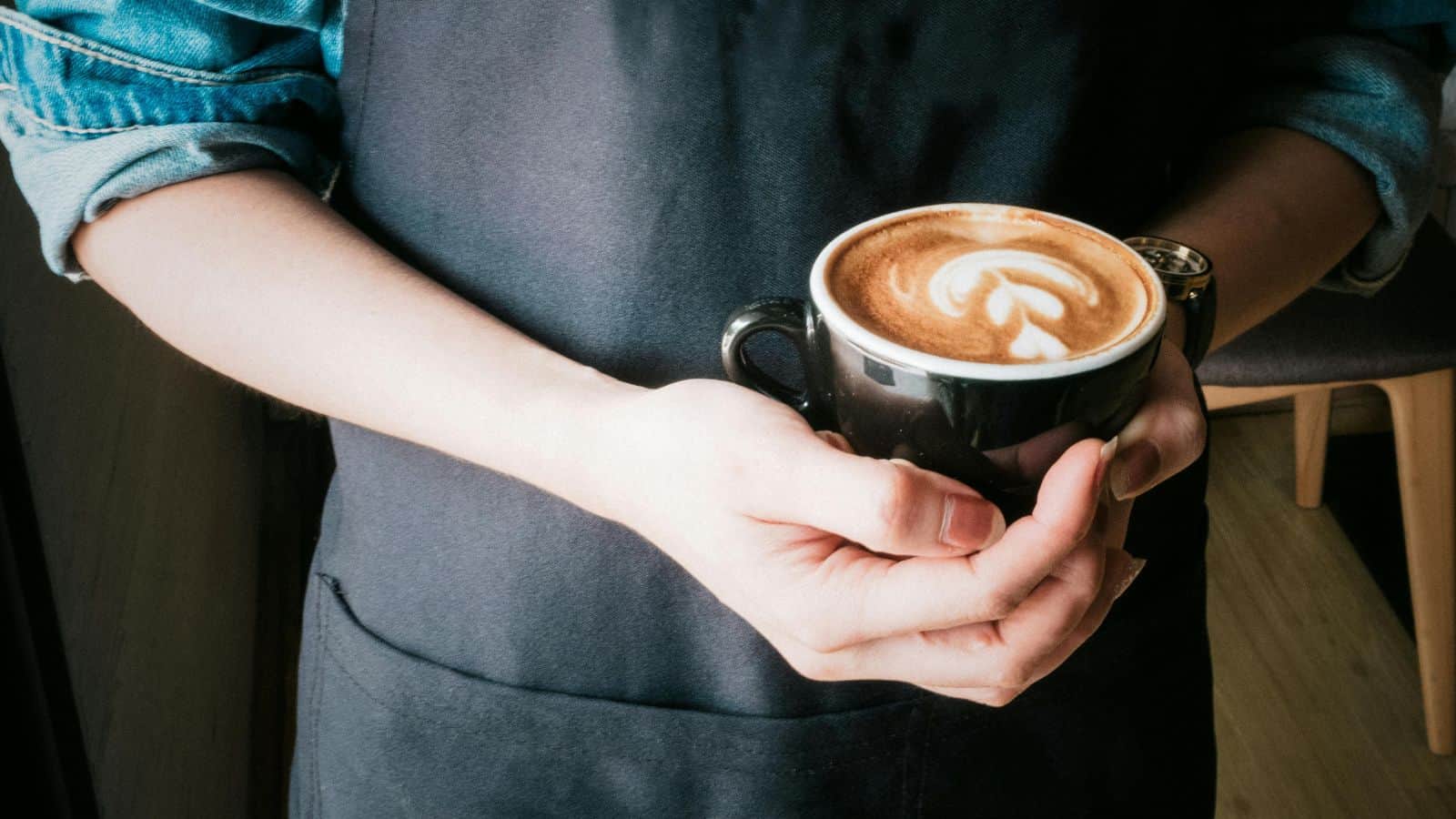 Person in a dark apron holding a cup of coffee with latte art on top, ready to be your guide to different types of coffee.