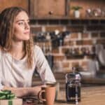 Woman with long hair sits at a kitchen counter with coffee, a French press, and a gift box in front of her, thoughtfully choosing between French press and pour over coffee for her morning brew.
