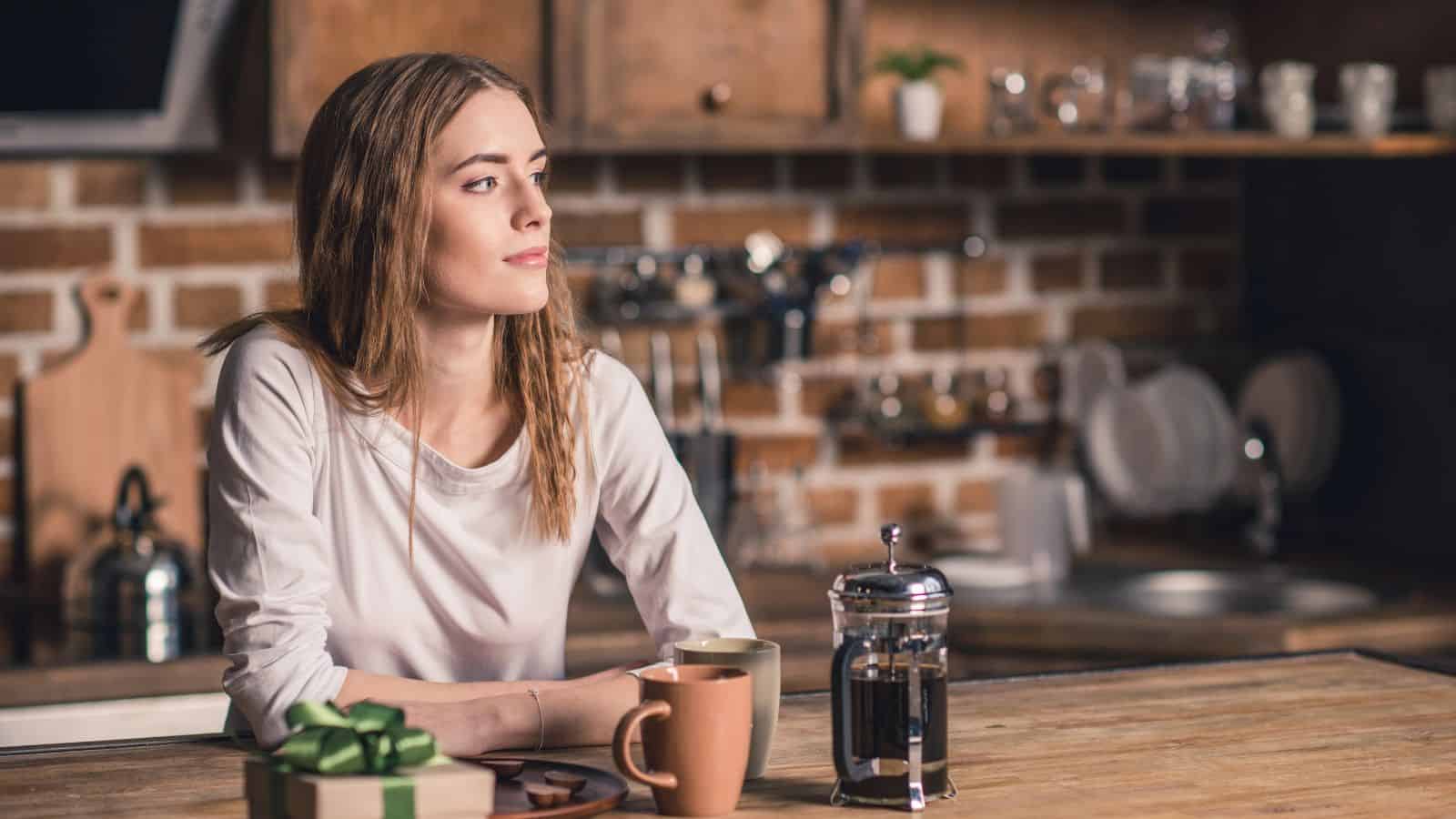 Woman with long hair sits at a kitchen counter with coffee, a French press, and a gift box in front of her, thoughtfully choosing between French press and pour over coffee for her morning brew.