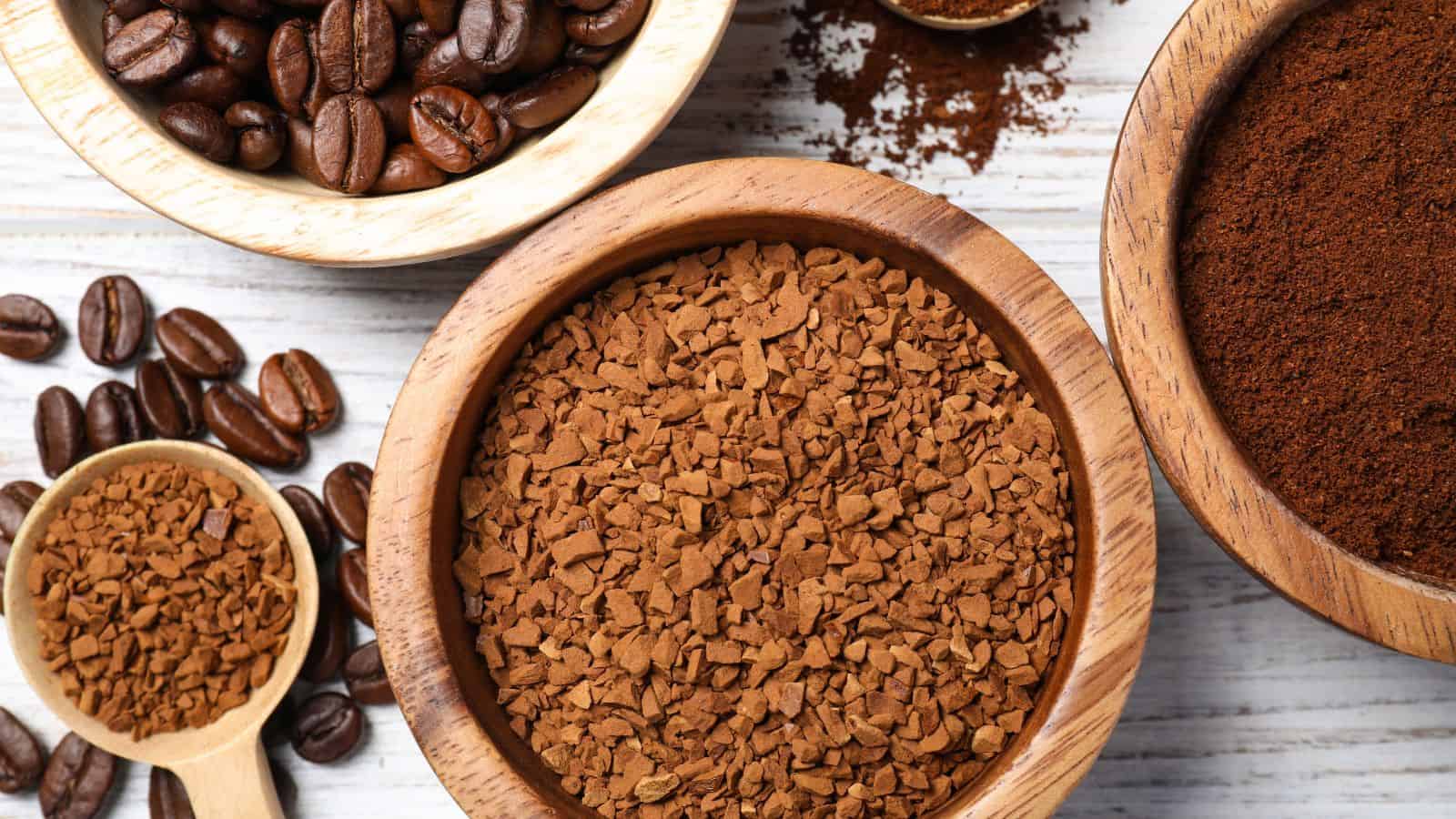 Bowls of instant coffee granules, ground coffee, and coffee beans on a white wooden surface—an inviting guide to different types of coffee.
