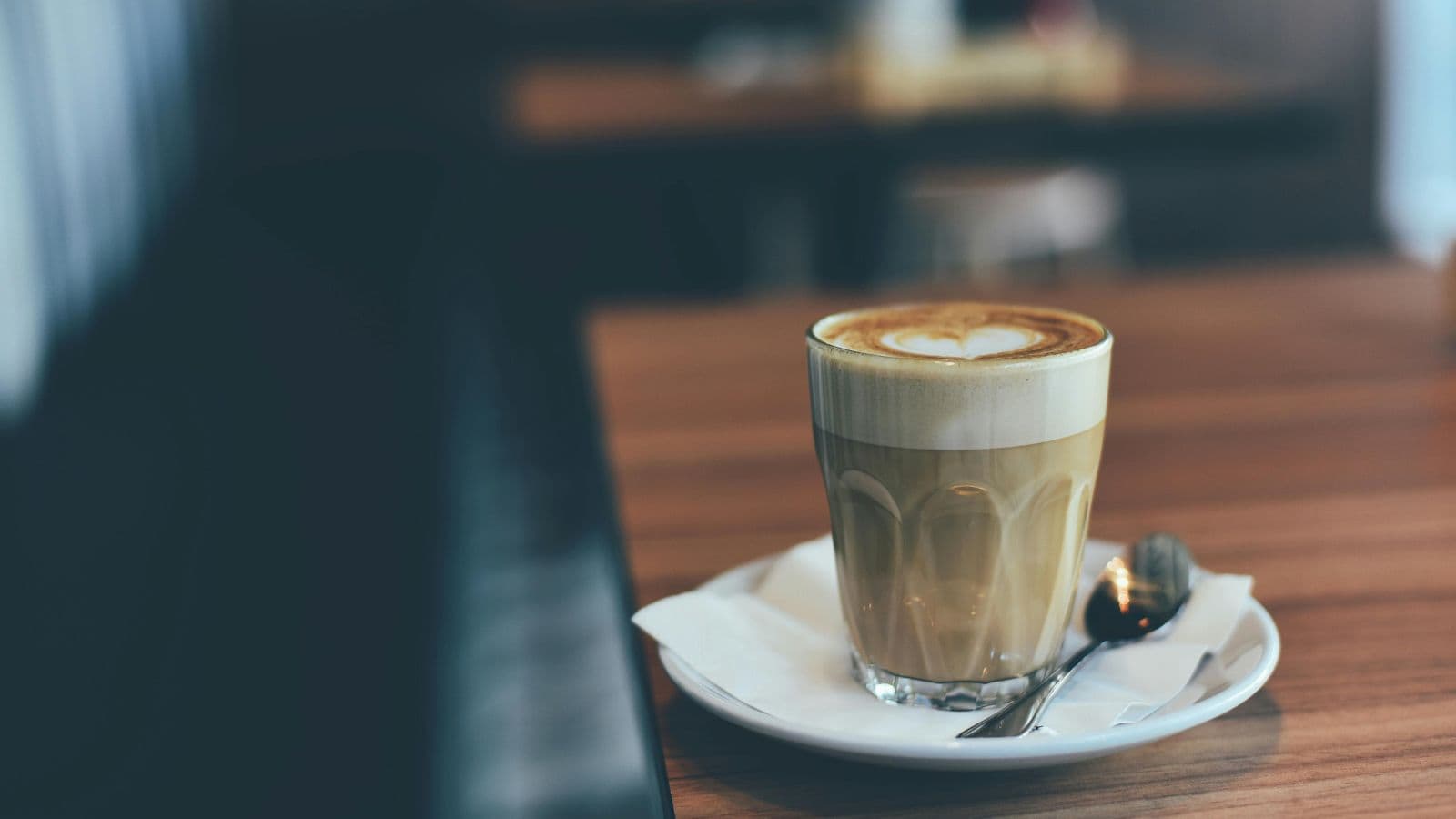 A glass of latte with heart-shaped foam sits on a saucer with a spoon on a wooden table in a café, perfect for anyone exploring a guide to different types of coffee.
