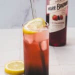 A tall glass of pink 4th of July lemonade with a lemon slice, straw, lemon wedges, and a grenadine bottle in the background.