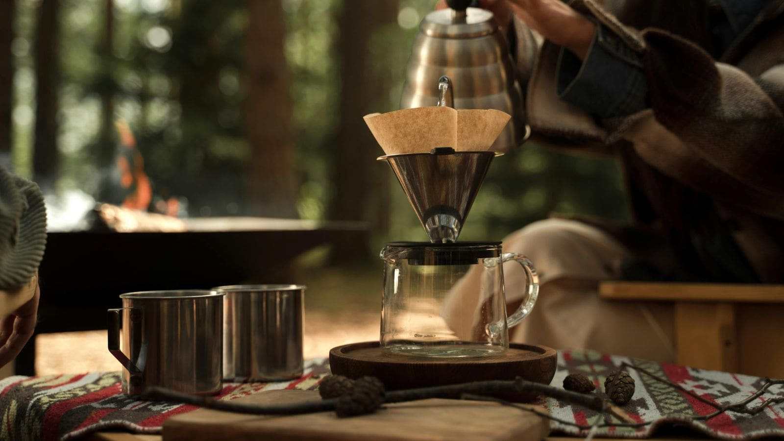 Pour-over coffee being made at a campsite, with mugs and a fire pit in the background, sets the scene for choosing between French press and pour over coffee on your next outdoor adventure.