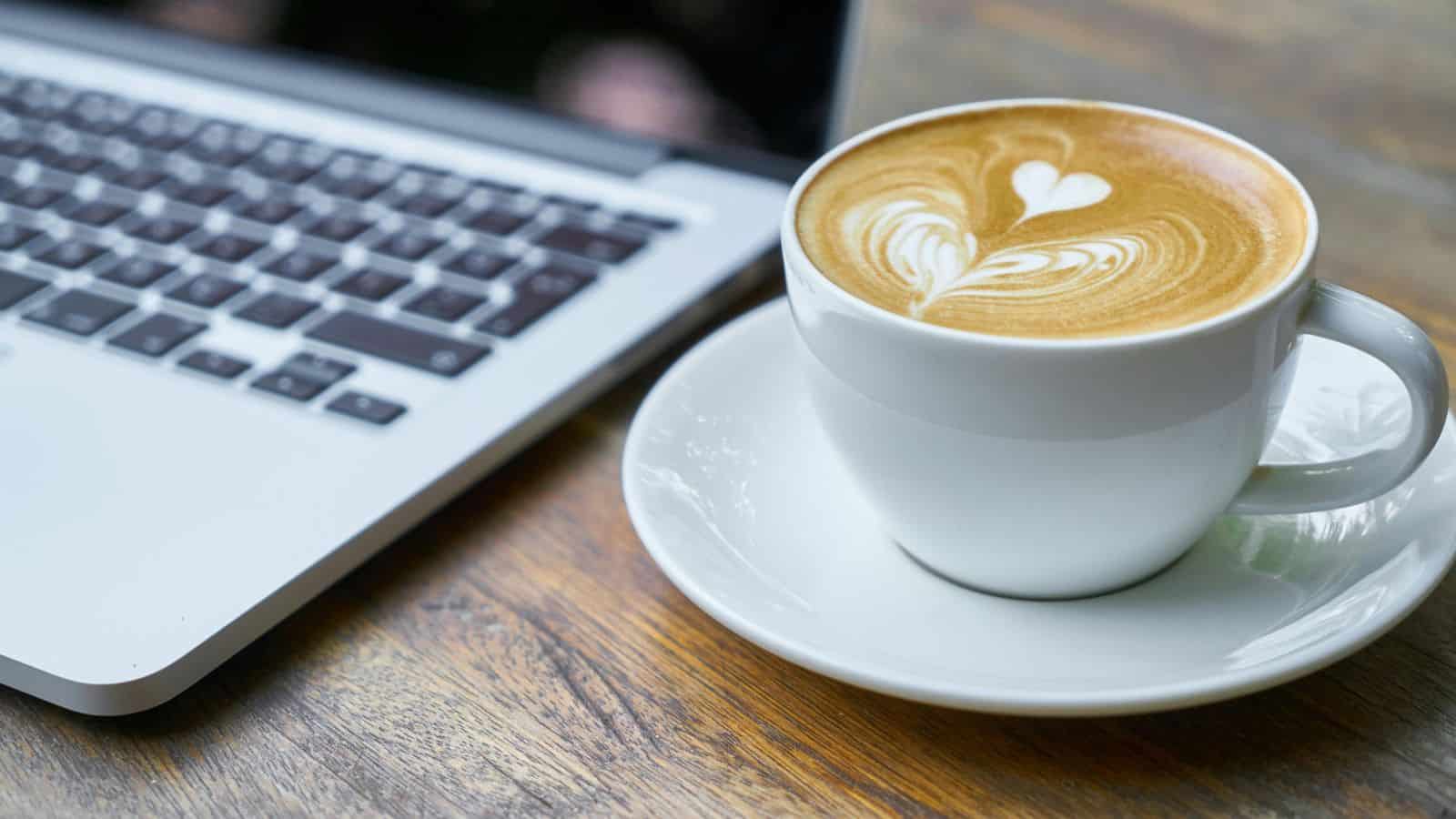 A cup of latte with heart latte art sits next to a laptop on a wooden table, perfect for reading a guide to different types of coffee.