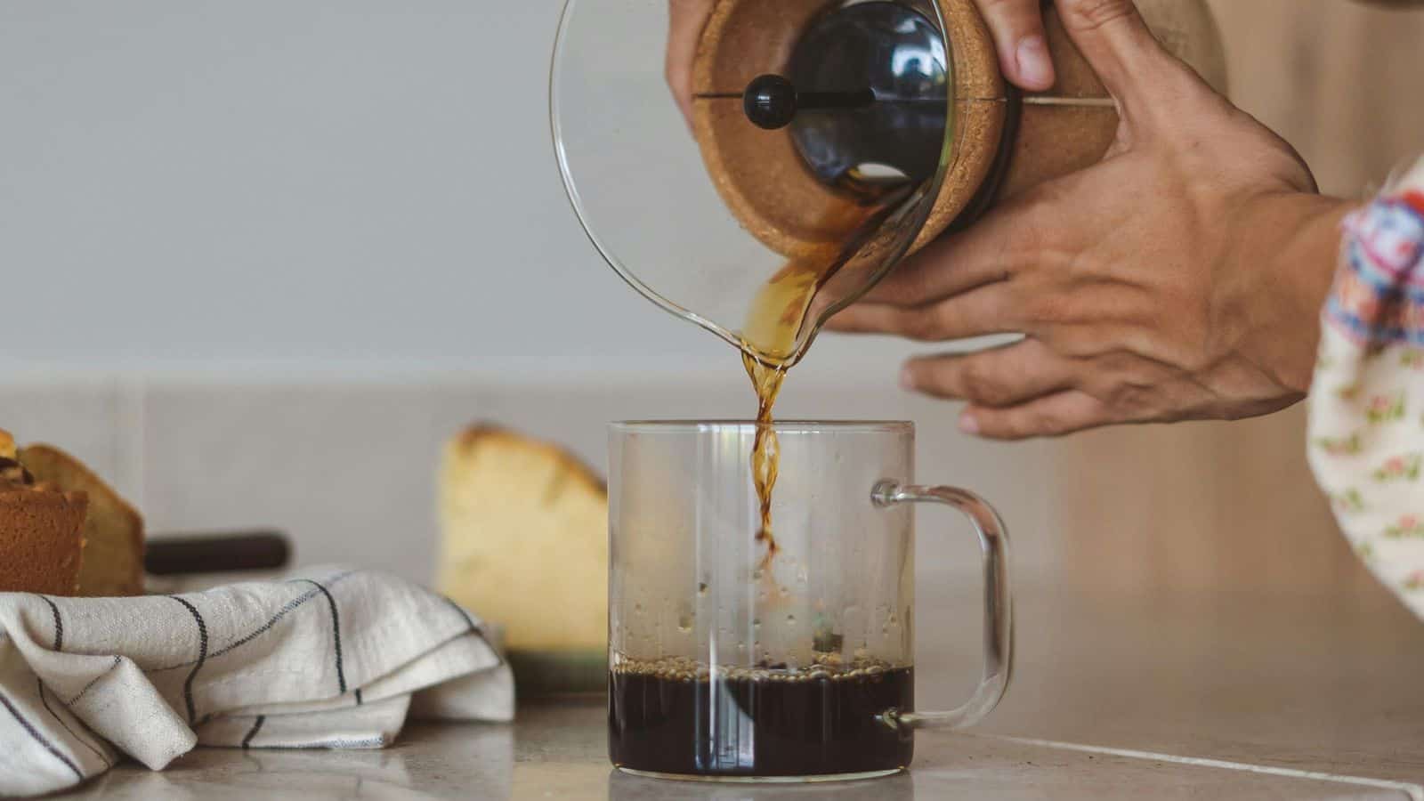 A person pours coffee from a French press into a glass mug on a kitchen counter, illustrating the ritual of choosing between French press and pour over coffee for the perfect morning brew.