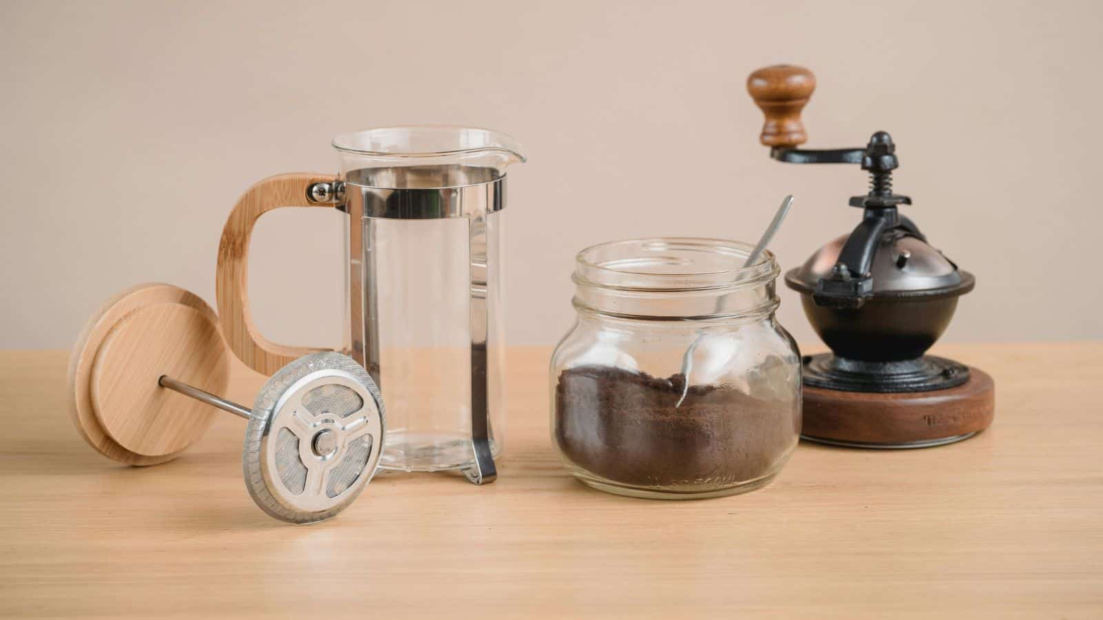 A French press, a jar of ground coffee with a spoon, and a manual coffee grinder on a wooden table—perfect aids for choosing between French press and pour over coffee.