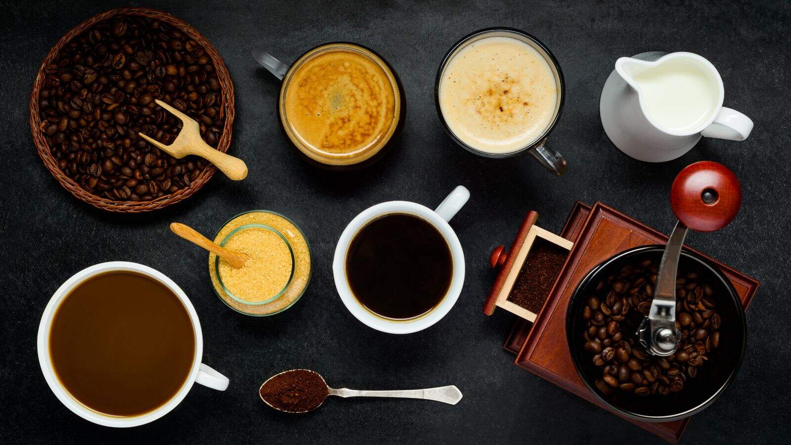 Top view of various coffee drinks, coffee beans, ground coffee, and a milk jug on a dark surface—a perfect guide to different types of coffee.