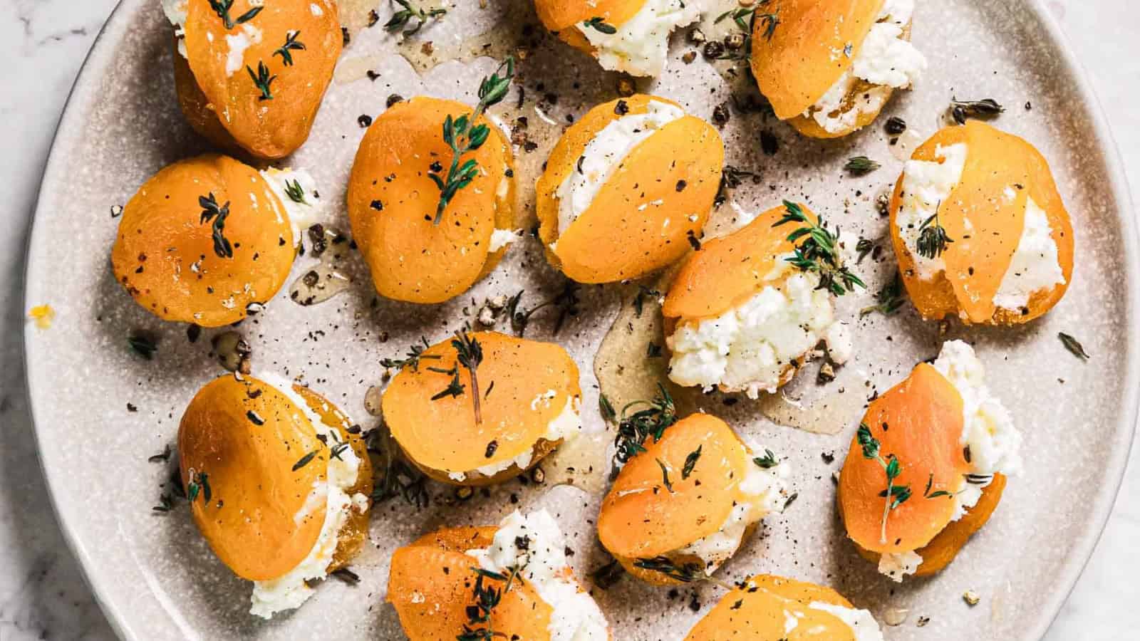 A plate of halved dried apricots stuffed with white cheese, garnished with fresh thyme and cracked black pepper, arranged on a round ceramic plate.