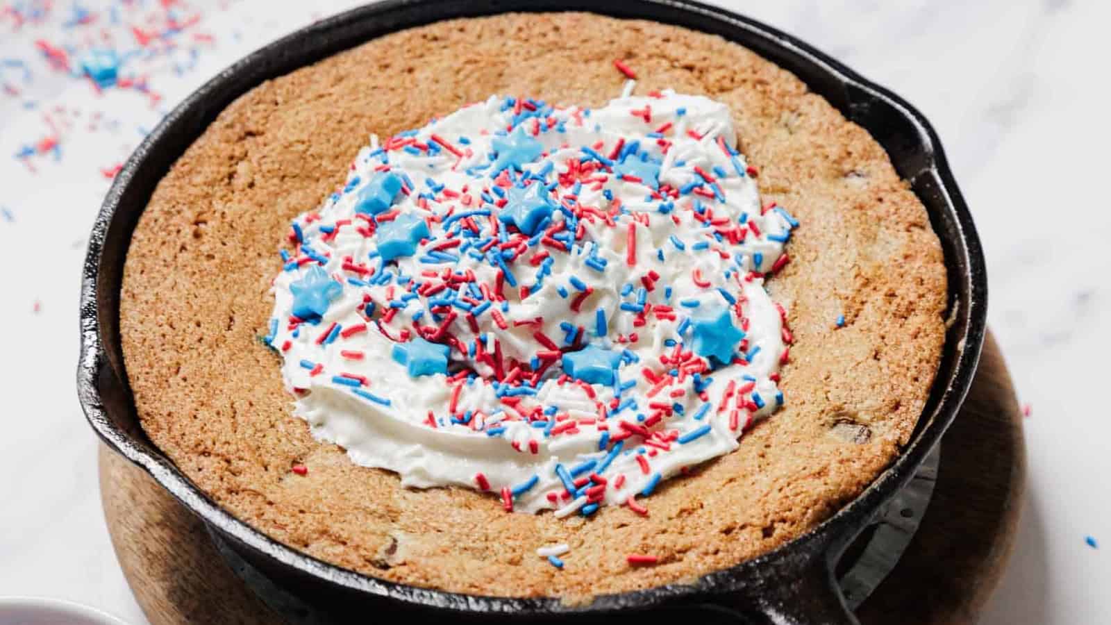 A large chocolate chip cookie baked in a cast iron skillet, topped with white frosting and red, white, and blue sprinkles, viewed from above on a white surface.