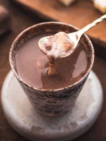 A spoon holding a thick, creamy chocolate mixture hovers over a cup filled with hot chocolate, set on a coaster on a wooden table. Chocolate blocks and a cutting board are visible in the background.