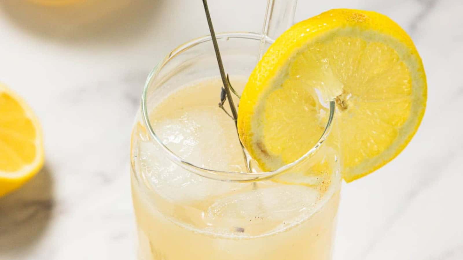 A glass and a carafe filled with lemonade are on a marble surface. The glass is garnished with a lemon slice and a sprig of lavender. Whole and halved lemons and a lavender sprig are scattered around.