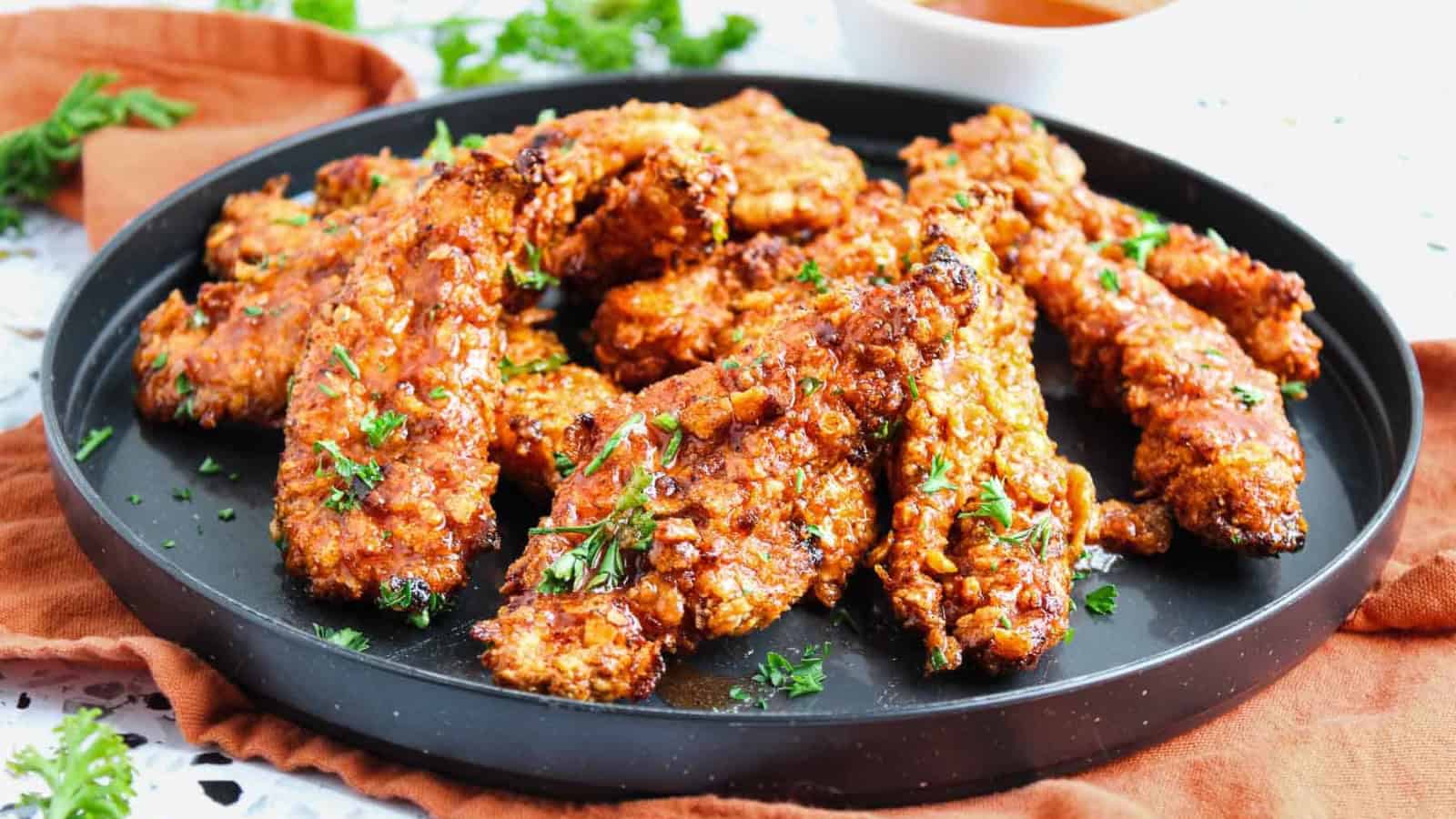 A black plate holding several pieces of golden-brown, crispy fried chicken tenders garnished with chopped parsley, set on a rust-colored cloth with sauce and herbs visible in the background.