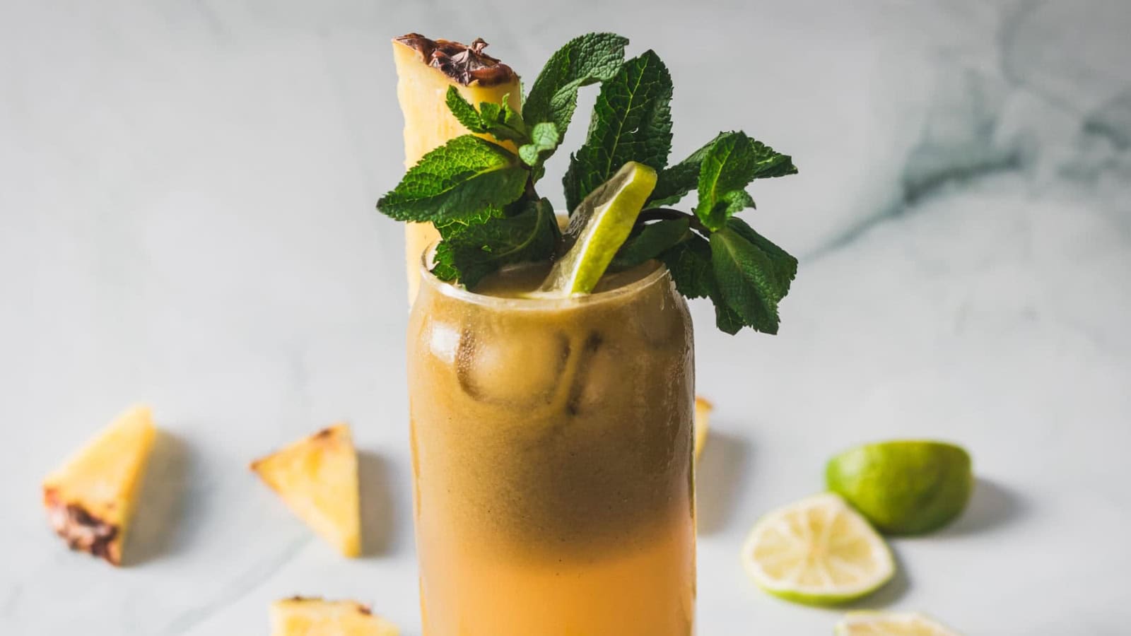 A tall glass filled with a light brown iced drink, garnished with a pineapple wedge, a lime slice, and a sprig of mint. Slices of lime and pineapple pieces are scattered on the white surface in the background.