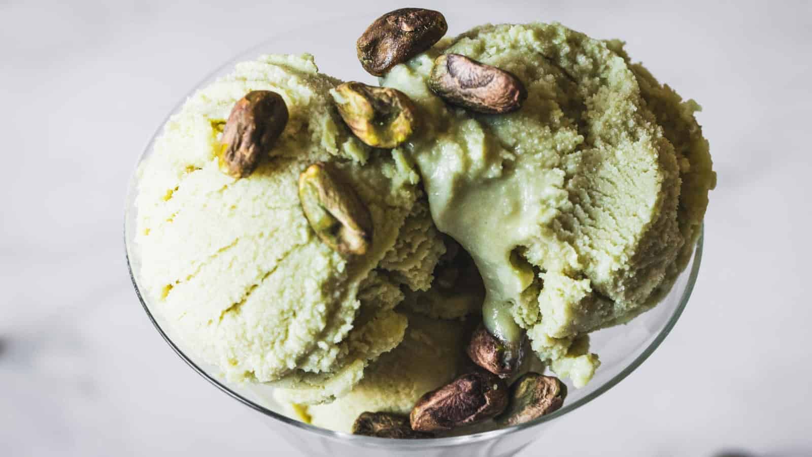 Three scoops of pistachio ice cream served in a glass dish, topped with several whole pistachio nuts, against a blurred light background.