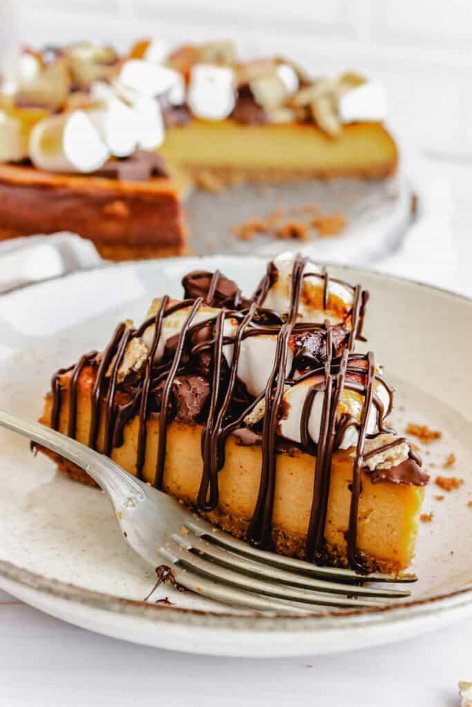 A slice of pumpkin smores cheesecake topped with marshmallows, chocolate, and graham cracker pieces, drizzled with chocolate.