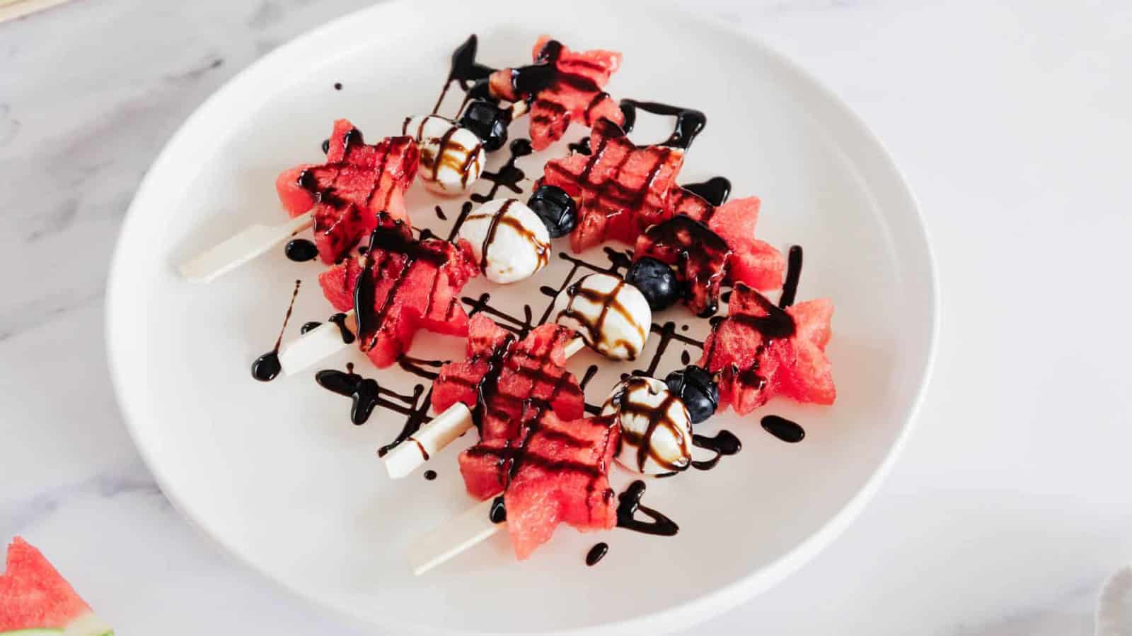 Watermelon and blueberry skewers with mozzarella balls are arranged on a white plate, drizzled with balsamic glaze. The skewers are placed on a marble surface.