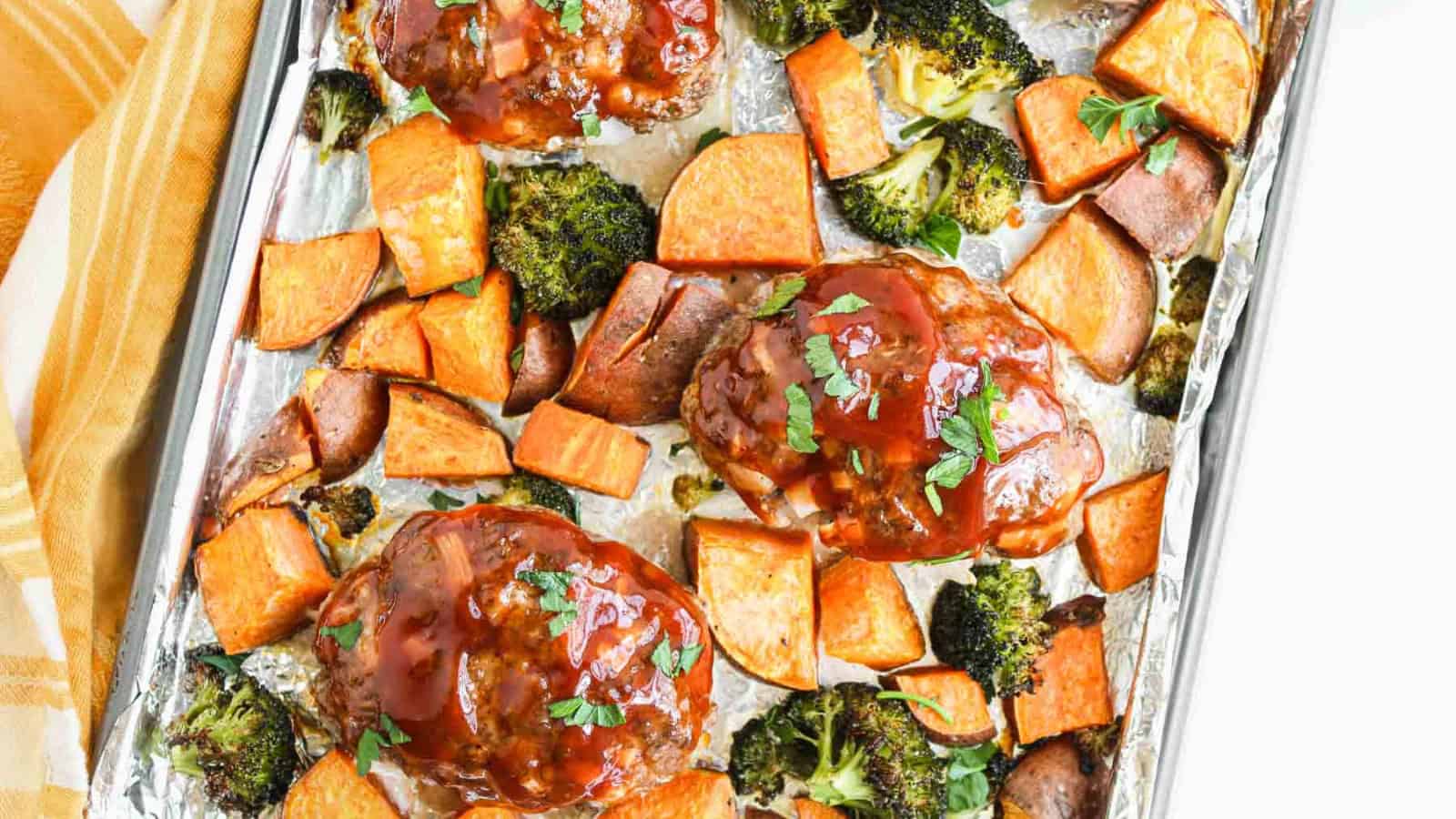 A plate with meatloaf topped with brown sauce, surrounded by roasted broccoli, sweet potato chunks, and roasted potatoes. A yellow-striped cloth is partially visible to the right of the plate.