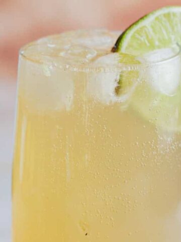 A tall glass filled with a light yellow fizzy drink, ice cubes, and a lime wheel garnish on the rim. A blurred bottle is visible in the background.