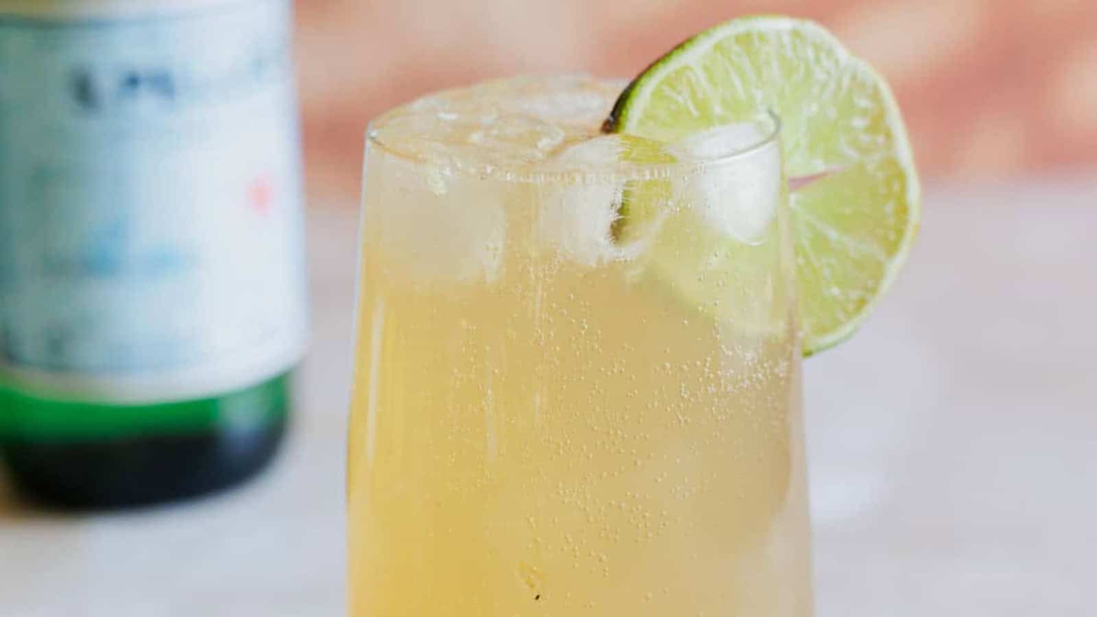 A tall glass filled with a light yellow fizzy drink, ice cubes, and a lime wheel garnish on the rim. A blurred bottle is visible in the background.