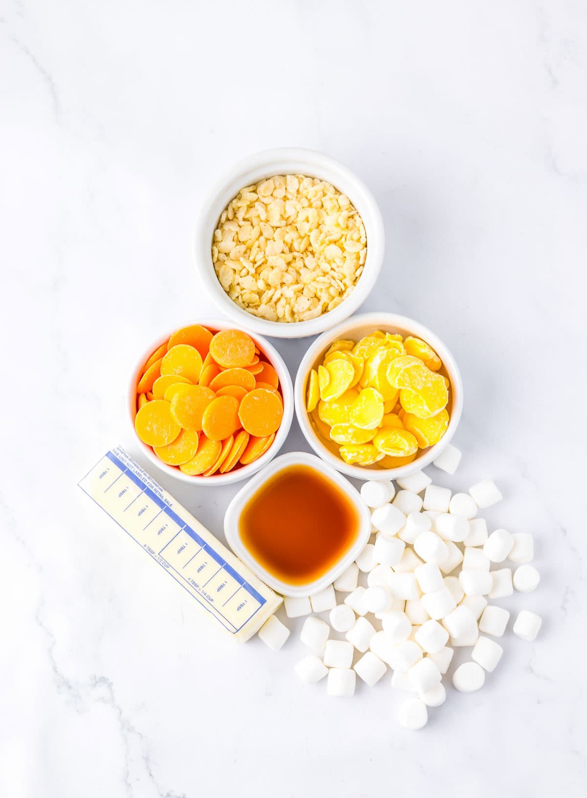 Ingredients for candy corn Rice Krispies&mdash;including mini marshmallows, butter, vanilla, and yellow candy melts&mdash;are arranged in bowls on a white surface.