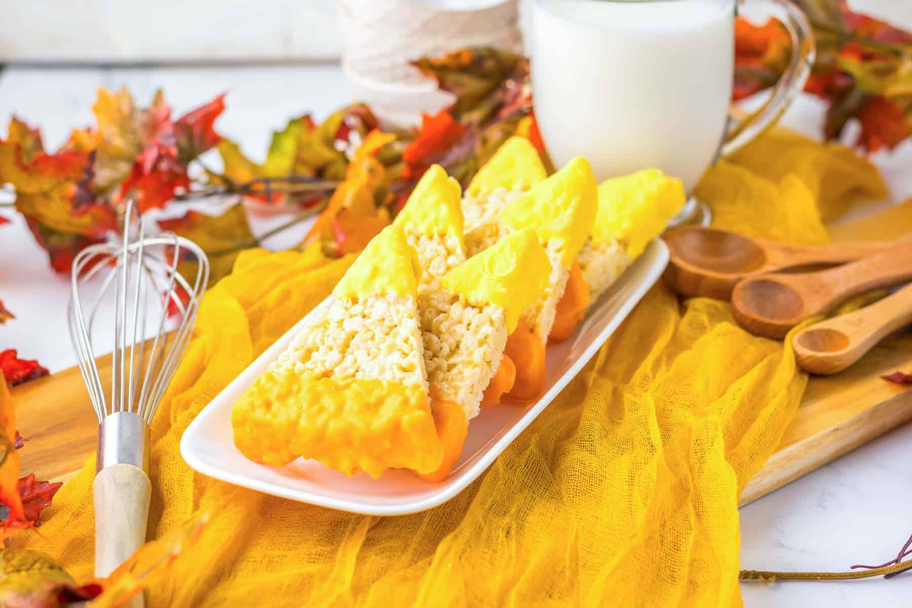 Three candy corn rice krispies treats sit on a plate with a glass of milk, surrounded by autumn leaves in the background.
