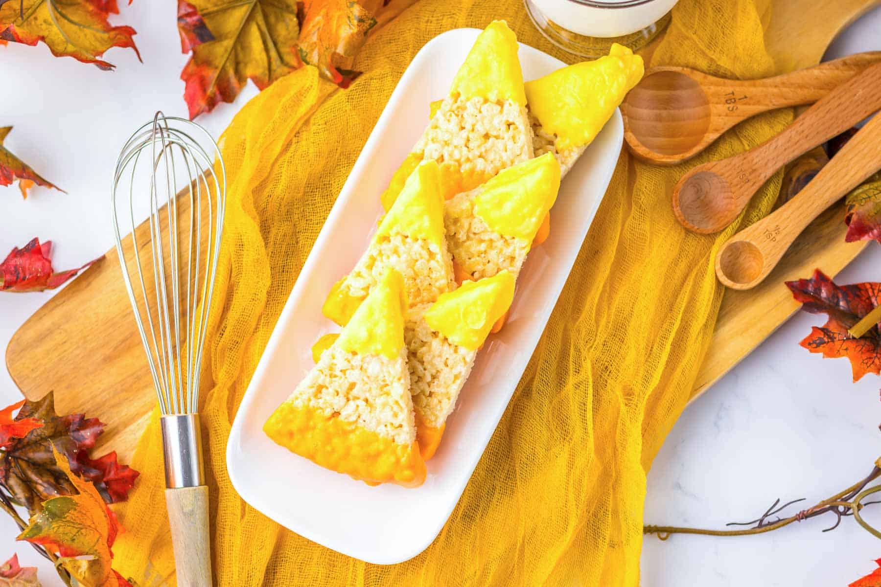 Candy corn Rice Krispies treats arranged on a white plate, surrounded by festive fall-themed items, create a sweet seasonal display.
