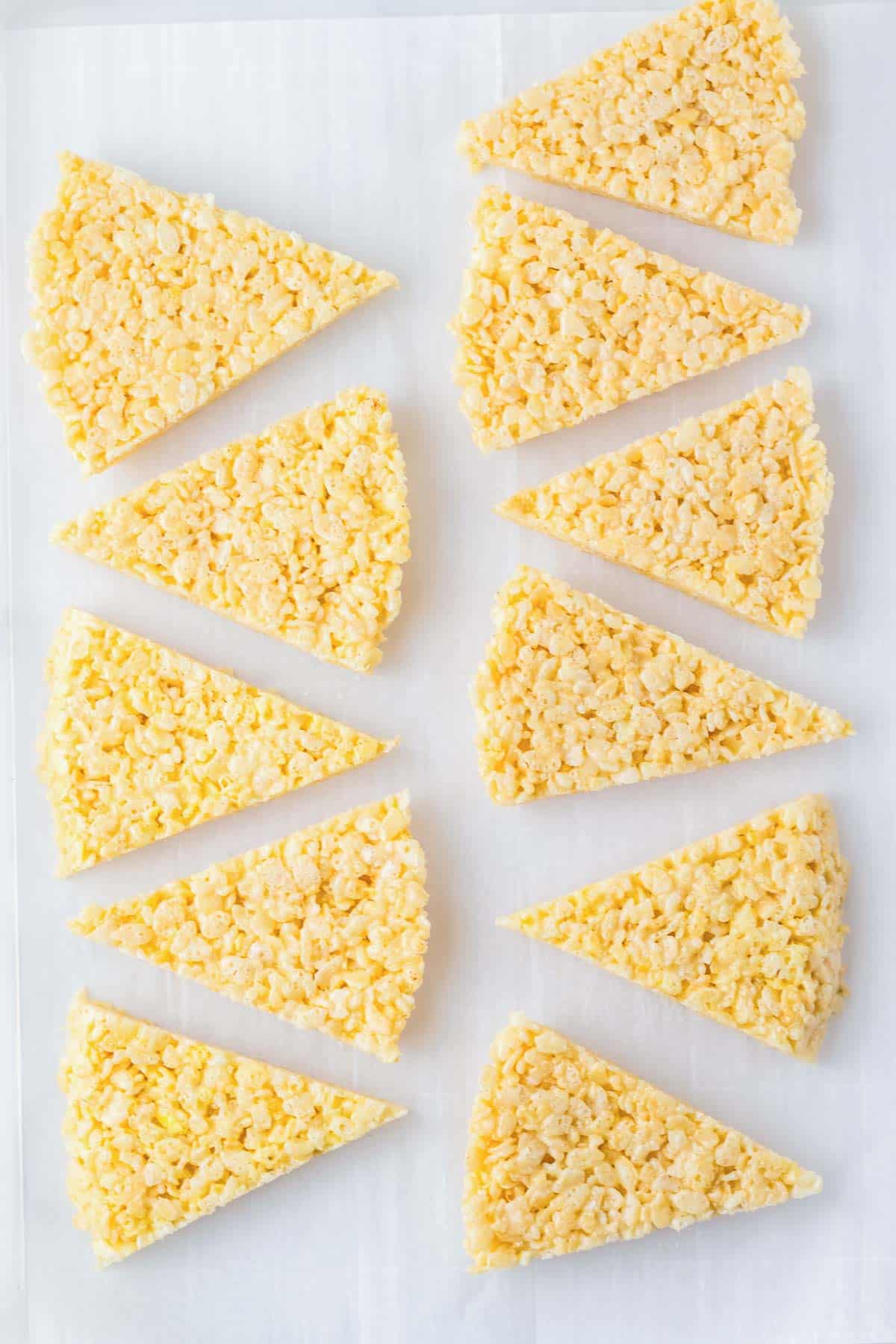 Twelve candy corn rice krispies treats, shaped into triangles, are arranged in neat rows on a white surface.