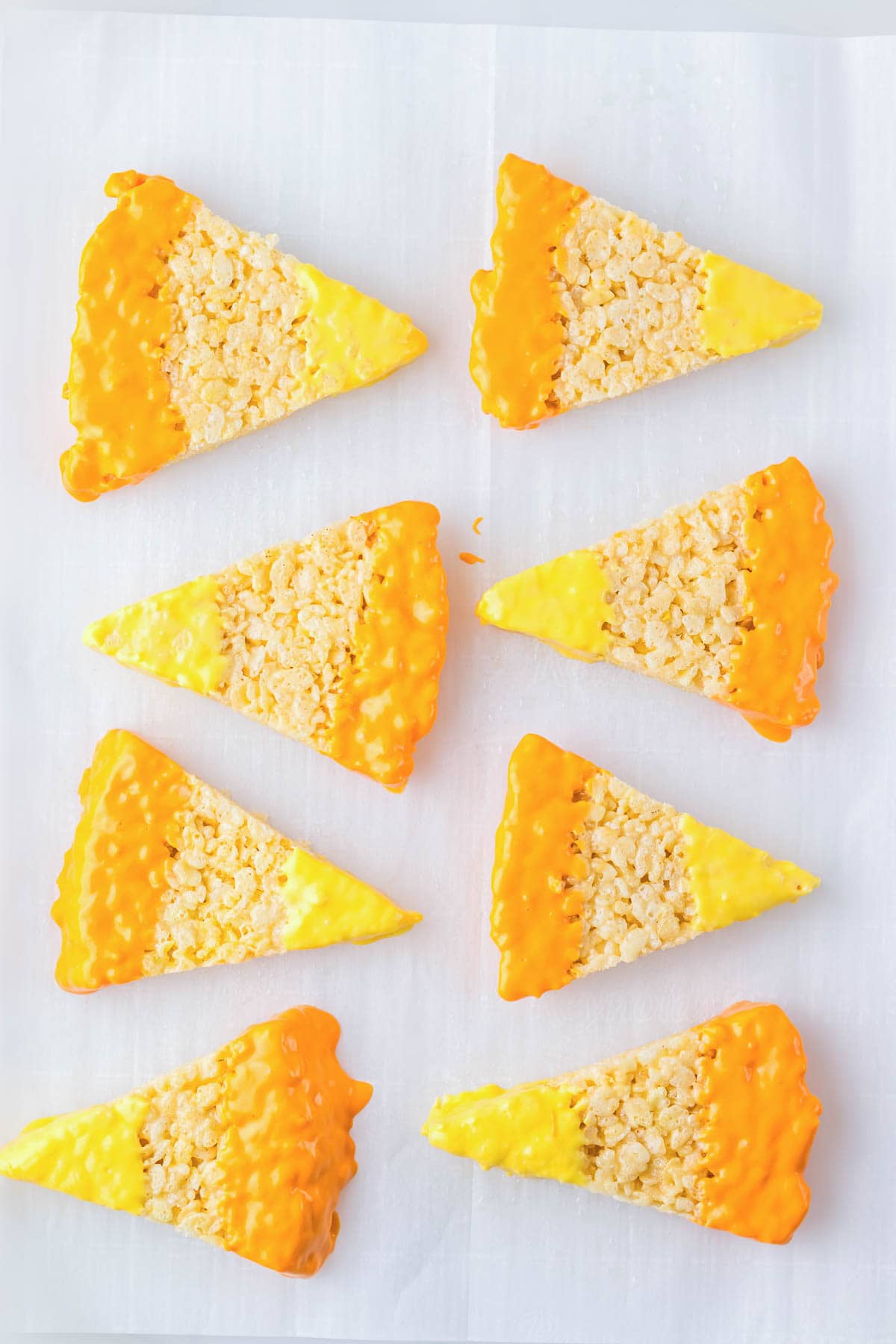Eight candy corn rice krispies shaped like classic treats, dipped in yellow and orange icing, arranged on white parchment paper.