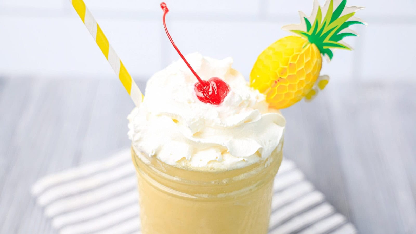 A creamy yellow drink topped with whipped cream, a cherry, a striped straw, and a paper pineapple decoration.