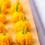 Mini pumpkin cheese balls shaped like pumpkins, with pretzel sticks and parsley as stems, are arranged on a parchment-lined tray.