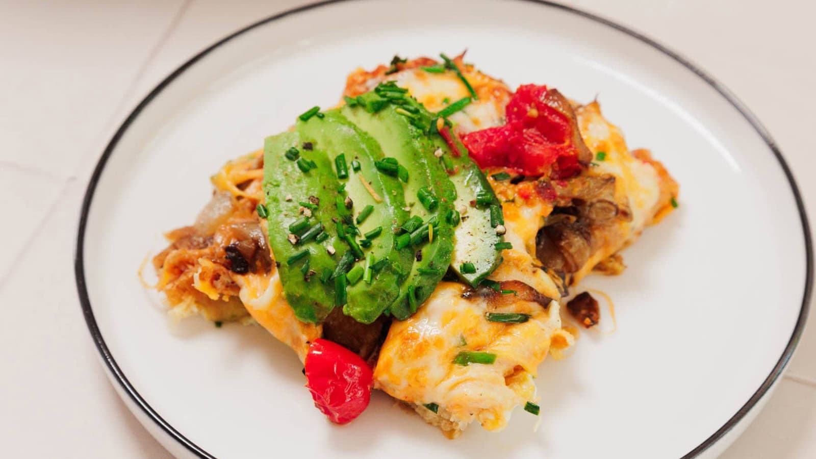 A serving of cheesy enchiladas topped with sliced avocado, chives, and cherry tomatoes on a white plate.