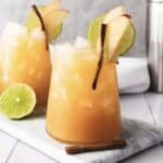 Iced apple cider cocktail with lime slices, apple slices, and a cinnamon stick garnish on a marble board&mdash;refreshing as a mocktail or with a splash of ginger beer.